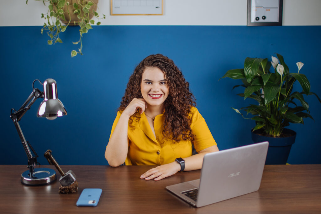 dívidas-em-dia: Veridiana Lopes, educadora financeira, de blusa amarela sentada em uma mesa e de frente para o notebook sorrindo