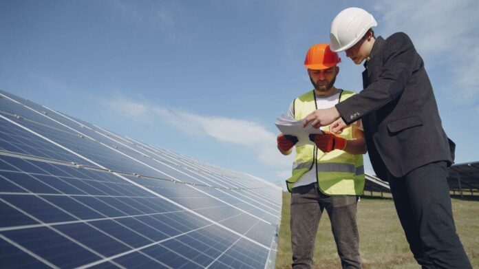 Dois homens de capacete de segurança analisam um projeto de instalação de energia solar em frente a um painel solar