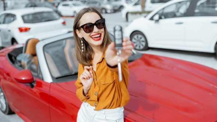 Mulher segura chave de carro e sorri, sentada no capô de um veículo vermelho