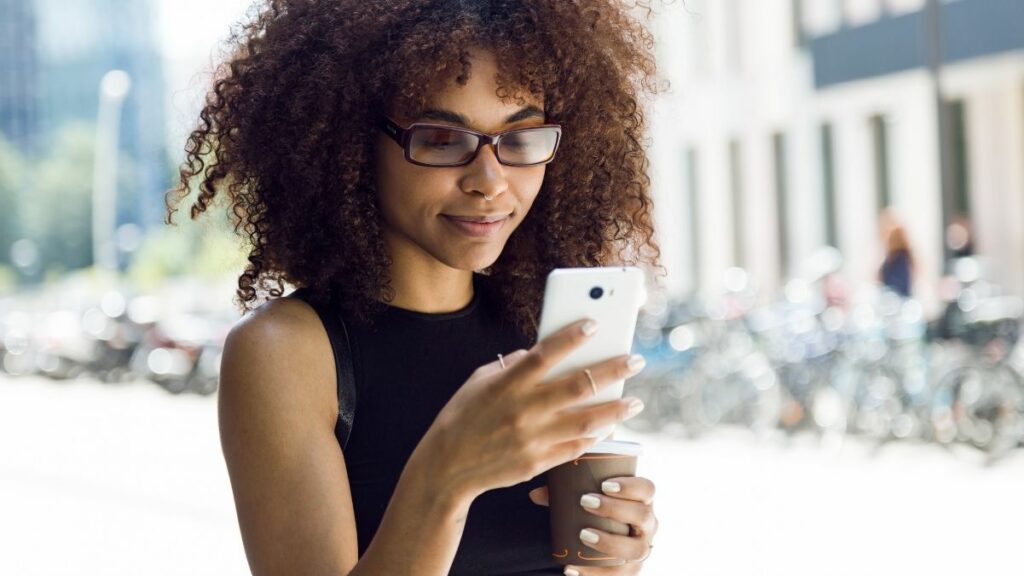 Menina com blusa preta segura celular nas mãos e olha para a tela sorrindo