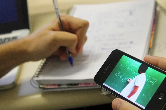 estudante escrevendo em um caderno e com a outra mão segurando o celular que passa um aula