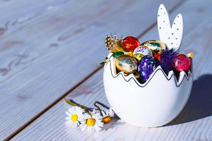 Recipiente branco em formato de ovo e com orelhas de coelho cheio de bombons e com flores ao lado